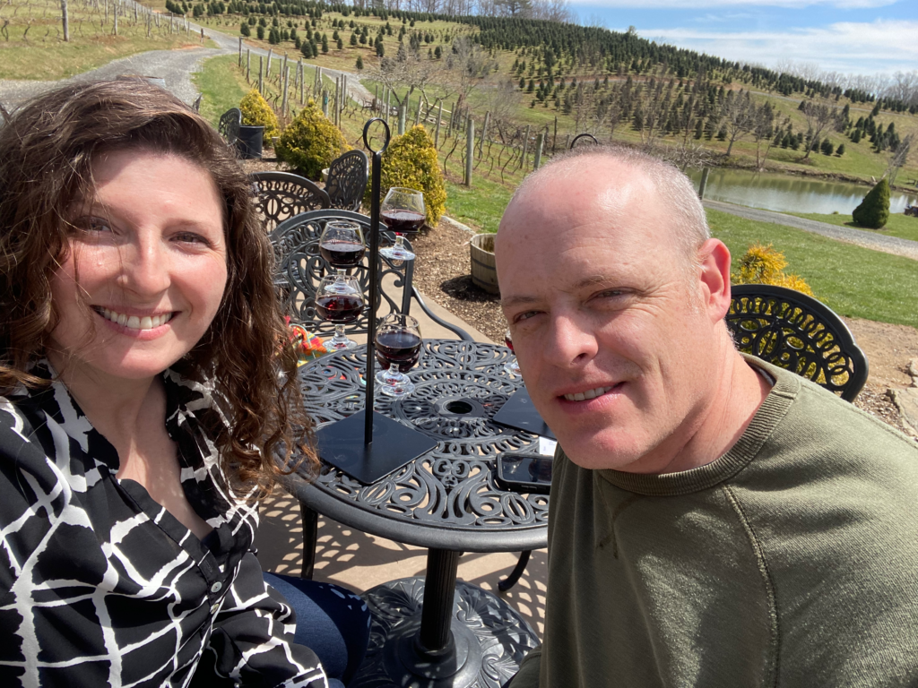 Photo of Marcianne O'Day and Buz Lee. They are sitting at a patio table with a vineyard in the background.